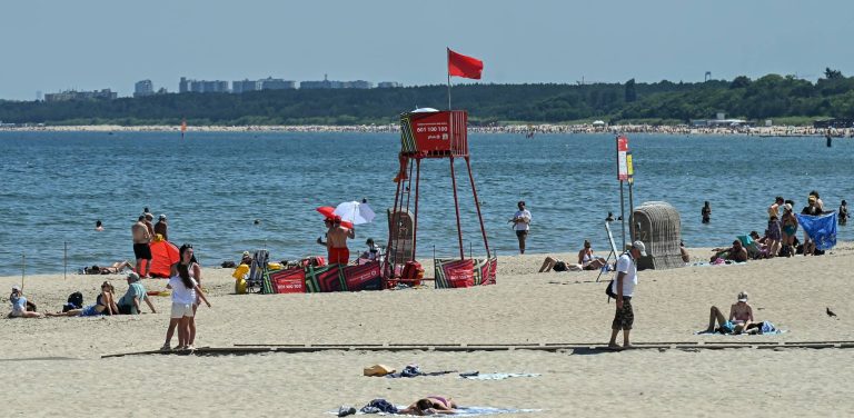 Słoneczny dzień na piaszczystej plaży nad morzem. W centralnej części kadru stoi czerwona, metalowa wieżyczka ratownicza, na której szczycie powiewa czerwona flaga oznaczająca zakaz kąpieli (tym razem z powodu sinic) Na plaży widoczni są wypoczywający na kocach ludzie oraz pojedyncze osoby spacerujące po drewnianej ścieżce wzdłuż piasku. W tle rozciąga się spokojna woda Zatoki Gdańskiej, a na horyzoncie widać zieloną linię drzew oraz zarysy miejskiej zabudowy