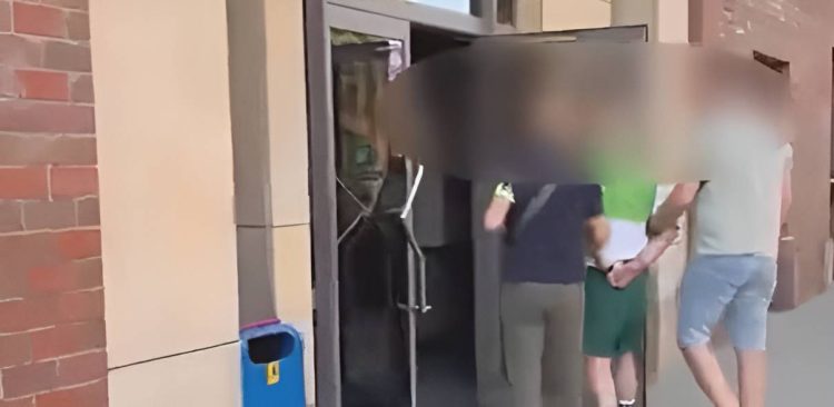 Zatrzymanie podejrzanego o groźby i znieważenie na tle narodowościowym obywatelki Ukrainy pseudokibica z Trójmiasta. Trzech mężczyzn stoi tyłem do obiektywu; środkowy z nich, ubrany w zieloną koszulkę i ciemne spodenki, ma ręce spięte z tyłu i jest prowadzony przez dwóch pozostałych mężczyzn. Twarze wszystkich osób na zdjęciu zostały mocno rozmyte w celu ochrony tożsamości. Po lewej stronie widoczny jest fragment ceglanej ściany oraz niebieski kosz na śmieci.
