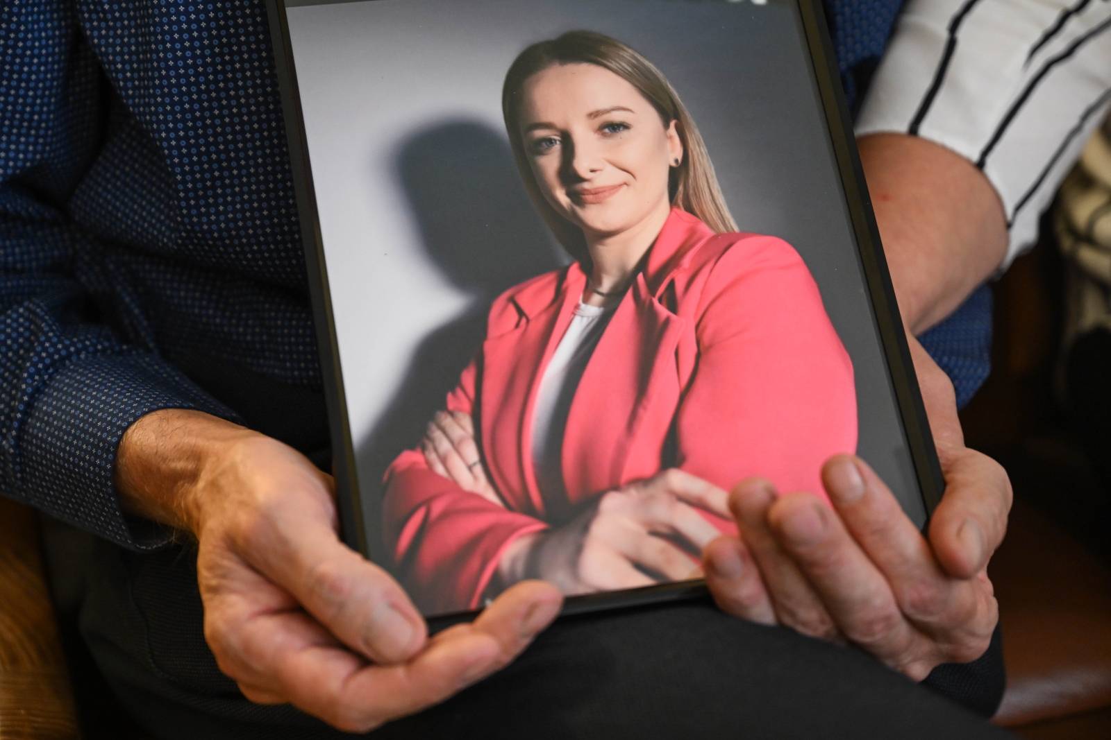 Dwie osoby trzymają w dłoniach oprawiony w ramkę portret uśmiechniętej młodej kobiety o blond włosach, ubranej w różową marynarkę. To zdjęcie kobiety najprawdopodobniej zamordowanej przez męża, który potem upozorował wypadek kolejowy w Mezowie na Kaszubach 