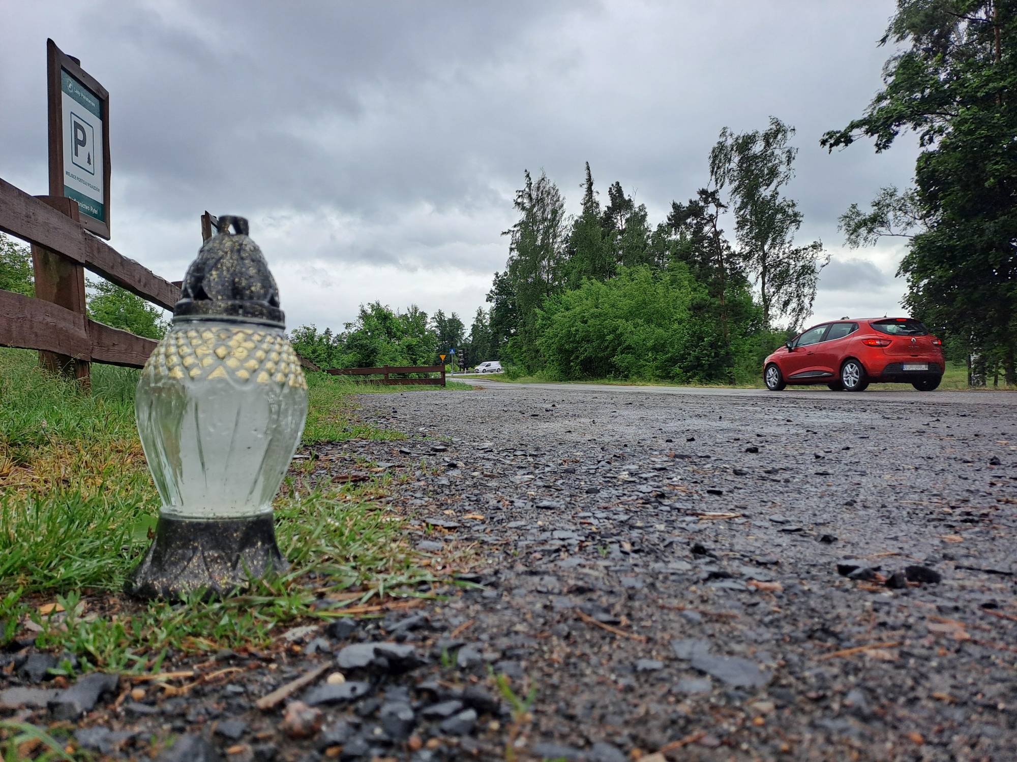 Znicz na parkingu obok wiejskiej drogi w gminie Czersk, gdzie zamordowano taksówkarza. Znicz po lewej stronie, nad nim płot drewniany oraz tablica informacyjna "P" (parking), po prawej stronie droga, na niej czerwony samochód, w oddali na zakręcie widać kolejne auto.