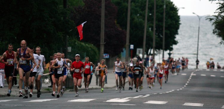 Nocny Bieg Świętojański. Zdjęcie przedstawia dużą grupę biegaczy uczestniczących w ulicznym biegu w Gdyni, prawdopodobnie maratonie lub półmaratonie. Biegacze są w różnym wieku, ubrani w sportowe stroje, a na ich klatkach piersiowych widoczne są numery startowe. Poruszają się po asfaltowej drodze, która lekko wznosi się w oddali i delikatnie zakręca w prawo. Po lewej stronie drogi widać zbocze porośnięte zielonymi krzewami i drzewami. Na tym zboczu znajduje się również słup, z którego powiewa czerwona flaga. W tle, po prawej stronie drogi, rozpościera się widok na morze lub duże jezioro, którego błękitna tafla łączy się z linią horyzontu. Wzdłuż drogi rozmieszczone są latarnie uliczne. Na asfalcie widoczne są białe linie, oddzielające pasy ruchu i wskazujące pobocze.