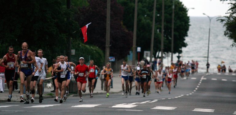 Nocny Bieg Świętojański. Zdjęcie przedstawia dużą grupę biegaczy uczestniczących w ulicznym biegu w Gdyni, prawdopodobnie maratonie lub półmaratonie. Biegacze są w różnym wieku, ubrani w sportowe stroje, a na ich klatkach piersiowych widoczne są numery startowe. Poruszają się po asfaltowej drodze, która lekko wznosi się w oddali i delikatnie zakręca w prawo. Po lewej stronie drogi widać zbocze porośnięte zielonymi krzewami i drzewami. Na tym zboczu znajduje się również słup, z którego powiewa czerwona flaga. W tle, po prawej stronie drogi, rozpościera się widok na morze lub duże jezioro, którego błękitna tafla łączy się z linią horyzontu. Wzdłuż drogi rozmieszczone są latarnie uliczne. Na asfalcie widoczne są białe linie, oddzielające pasy ruchu i wskazujące pobocze.