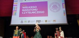 Scena teatralną lub aula, gdzie odbywa się wydarzenie. Na dużym ekranie w tle wyświetlany jest napis "Gala WIELKIEGO MARATONU CZYTELNICZEGO" oraz informacje o organizatorach i edycji 2024/2025. Pod ekranem, na scenie, stoi kilka osób w strojach stylizowanych. Centralną postacią jest kobieta w długiej, ciemnozielonej sukni z falbanami, która trzyma wysoko nad głową białą, ozdobną statuetkę. Po jej lewej stronie stoi osoba w stroju przypominającym mundurek wojskowy z epoki - w zielono-czerwone pasy i złotymi guzikami. Po prawej stronie stoi inna osoba w stroju w biało-czarne kropki. Na scenie widać również stosy książek ułożonych w niewielkie wieże. Oświetlenie sceny jest stonowane, publiczność jest poza kadrem.