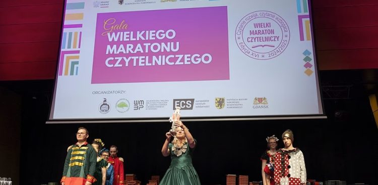 Scena teatralną lub aula, gdzie odbywa się wydarzenie. Na dużym ekranie w tle wyświetlany jest napis "Gala WIELKIEGO MARATONU CZYTELNICZEGO" oraz informacje o organizatorach i edycji 2024/2025. Pod ekranem, na scenie, stoi kilka osób w strojach stylizowanych. Centralną postacią jest kobieta w długiej, ciemnozielonej sukni z falbanami, która trzyma wysoko nad głową białą, ozdobną statuetkę. Po jej lewej stronie stoi osoba w stroju przypominającym mundurek wojskowy z epoki - w zielono-czerwone pasy i złotymi guzikami. Po prawej stronie stoi inna osoba w stroju w biało-czarne kropki. Na scenie widać również stosy książek ułożonych w niewielkie wieże. Oświetlenie sceny jest stonowane, publiczność jest poza kadrem.