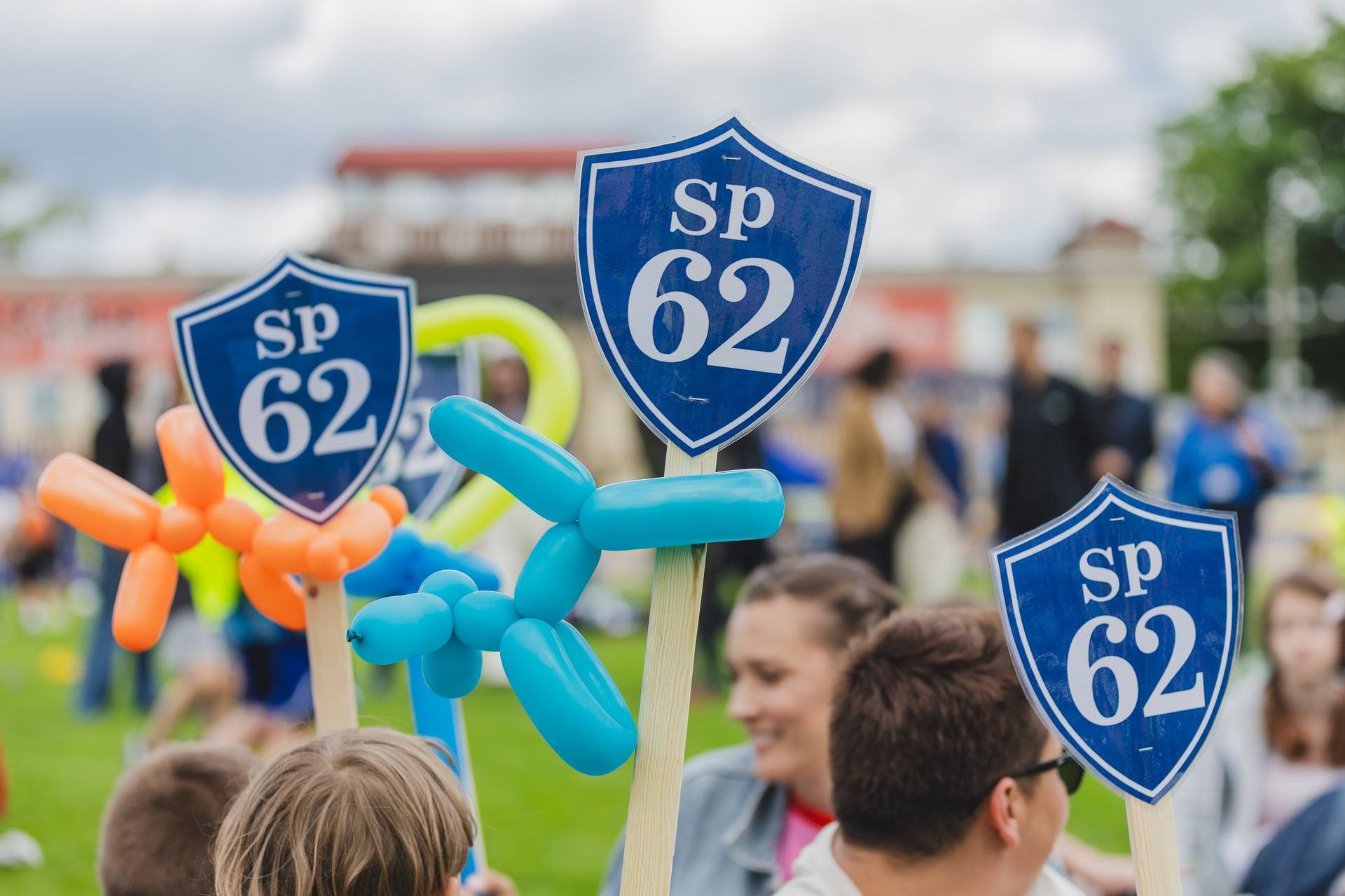 Uczestnicy Rowerowego Maja w Gdańsku podczas uroczystego zakończenie XII edycji akcji. Widać tabliczki z napisem "SP 62" oraz kolorowe balony w kształcie zwierząt. Uśmiechnięci ludzie na tle zielonego terenu