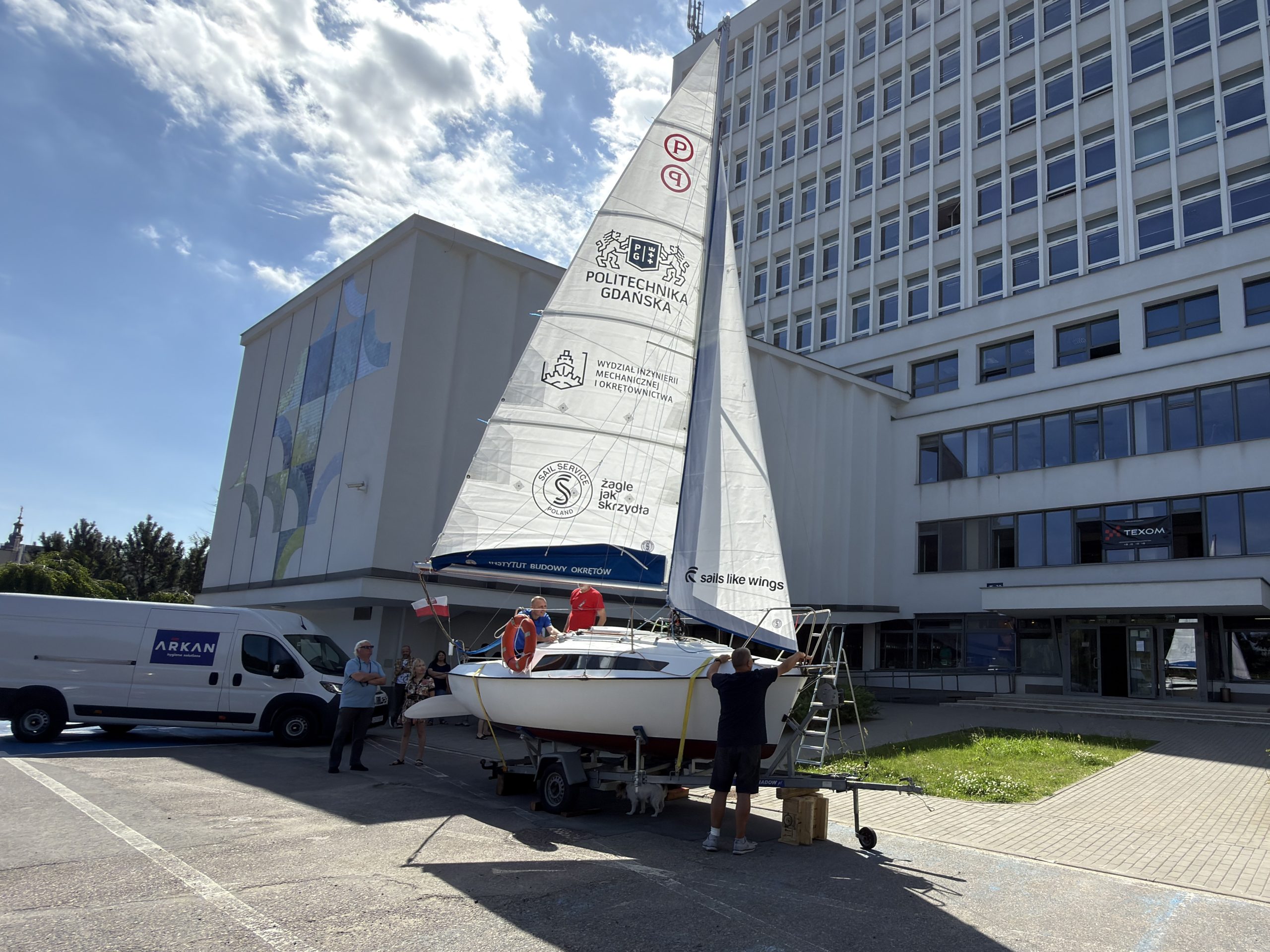 Zdjęcie przedstawia odnowiony jacht klasy Micro o nazwie „MicroPolo”, ustawiony na przyczepie transportowej na placu przed wielopiętrowym, jasnym budynkiem Politechniki Gdańskiej. Jacht ma postawione białe żagle (grot i fok). Na głównym żaglu widoczne są logotypy Politechniki Gdańskiej, Wydziału Inżynierii Mechanicznej i Okrętownictwa oraz firmy Sail Service. Na niebieskiej osłonie bomu znajduje się napis „Instytut Budowy Okrętów”. Przy jednostce pracuje kilka osób – jeden mężczyzna stoi przy dziobie, dwie osoby znajdują się w kokpicie, a z boku przyglądają się przechodnie. Po lewej stronie widoczny jest zaparkowany biały samochód dostawczy. Nad sceną rozciąga się częściowo zachmurzone, błękitne niebo