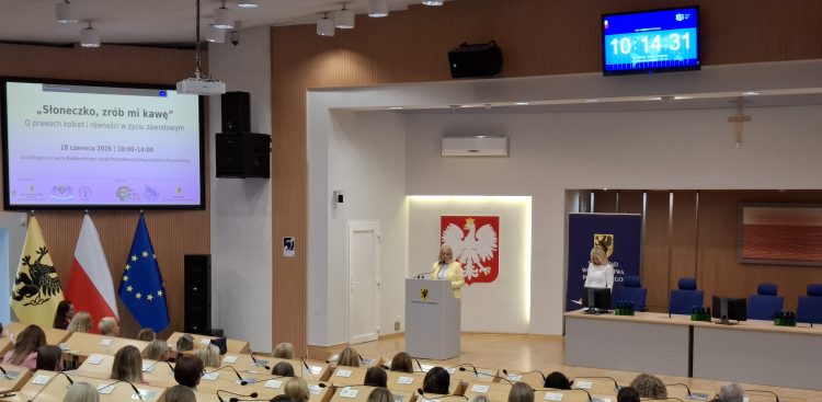 Sala w Urzędzie Marszałkowskim Województwa Pomorskiego. Odbywa się konferencja "Słoneczko, zrób mi kawę" dot. dyskryminacji kobiet w miejscu pracy. Na scenie, po lewej stronie, za mównicą stoi prelegentka. Na mównicy widoczny jest herb woj. pomorskiego (gryf), a za plecami prelegentki duże godło Polski (biały orzeł na czerwonym tle. Za sceną, na ścianie, wiszą flagi: Polski i i Unii Europejskiej. Po lewej stronie sceny znajduje się duży ekran projekcyjny, na którym wyświetlany jest slajd z tekstem w języku polskim, rozpoczynającym się od "Słoneczko, zrób mi kawę?" oraz informacjami o "Prawach kobiet i stereotypach w życiu zawodowym". W tle, po prawej stronie sceny, znajduje się zegar cyfrowy pokazujący godzinę 10:14:31. Obok zegara widać również drugą osobę, stojącą przy pulpicie. Pomieszczenie jest dobrze oświetlone
