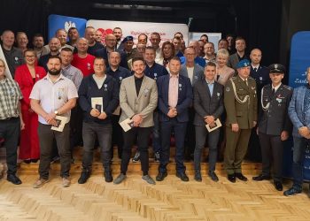 Uroczystość wręczenia medali „Honorowego Dawcy Krwi – Zasłużonego dla Zdrowia Narodu” w Słupsku. Na zdjęciu widać dużą grupę osób stojących w rzędach, z których większość to mężczyźni. Wszyscy ubrani są w eleganckie stroje, a wielu z nich ma przypięte do ubrań medale. Na pierwszym planie, po lewej stronie, stoi kilka osób z dokumentami w rękach, prawdopodobnie certyfikatami lub podziękowaniami. W tle widoczne są niebieskie banery z godłem Polski oraz napisy związane z krwiodawstwem. Całość ma charakter oficjalny i uroczysty