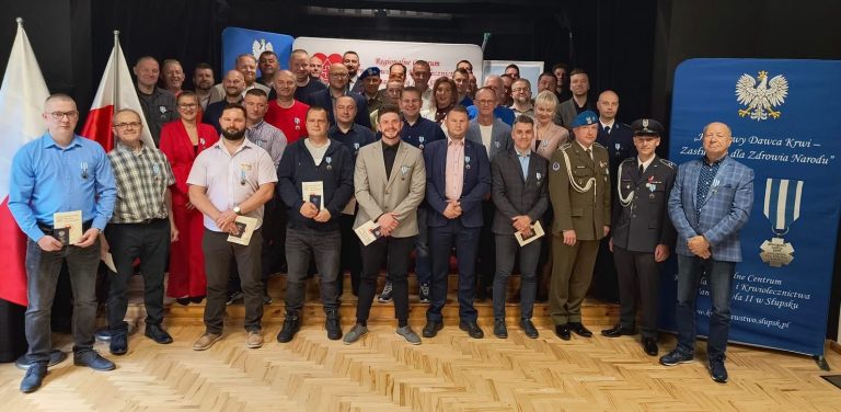 Uroczystość wręczenia medali „Honorowego Dawcy Krwi – Zasłużonego dla Zdrowia Narodu” w Słupsku. Na zdjęciu widać dużą grupę osób stojących w rzędach, z których większość to mężczyźni. Wszyscy ubrani są w eleganckie stroje, a wielu z nich ma przypięte do ubrań medale. Na pierwszym planie, po lewej stronie, stoi kilka osób z dokumentami w rękach, prawdopodobnie certyfikatami lub podziękowaniami. W tle widoczne są niebieskie banery z godłem Polski oraz napisy związane z krwiodawstwem. Całość ma charakter oficjalny i uroczysty