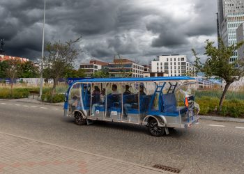 Niebiesko-srebrny, wieloosobowy pojazd elektryczny (darmowy meleks dla turystów w Gdyni) z pasażerami porusza się po ulicy w Gdyni. Pojazd ma zamontowane przezroczyste, foliowe osłony przeciwdeszczowe. W tle widoczne są miejskie tereny zielone, nowoczesna zabudowa oraz bardzo ciemne, burzowe chmury na niebie.
