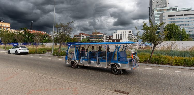 Niebiesko-srebrny, wieloosobowy pojazd elektryczny (darmowy meleks dla turystów w Gdyni) z pasażerami porusza się po ulicy w Gdyni. Pojazd ma zamontowane przezroczyste, foliowe osłony przeciwdeszczowe. W tle widoczne są miejskie tereny zielone, nowoczesna zabudowa oraz bardzo ciemne, burzowe chmury na niebie.