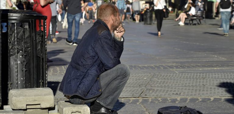 Mężczyzna w niebieskiej kurtce, prawdopodobnie osoba w kryzysie bezdomności, siedzi samotnie na betonowej płycie budowlanej mijany przez idących deptakiem ludzi - ilustruje potrzebę systemowego wsparcia, takiego jak wprowadzona przez Gdańsk Karta Praw Osób Bezdomnych