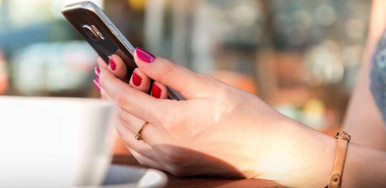 Telefon trzymany w kobiecych dłoniach, obok stoi filiżanka
