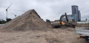 Plac budowy kompleksu Nova Marina w Gdyni. Na pierwszym planie, po lewej stronie, dominuje hałda piachu lub gruzu. Na prawo stoi żółta koparka gąsienicowa, która ładuje materiał na wywrotkę stojącą obok. Na ziemi widać ślady prac ziemnych i nieregularną, piaszczystą powierzchnię. W tle, po lewej stronie, widoczne są trzy żurawie budowlane, sugerujące dalsze prace konstrukcyjne w okolicy. Po prawej stronie, na horyzoncie, rysują się nowoczesne wieżowce, w tym jeden charakterystyczny, wysoki budynek o szklanej elewacji. Pomiędzy placem budowy a wieżowcami widać fragmenty innych budynków i miejskiej infrastruktury i zachmurzone