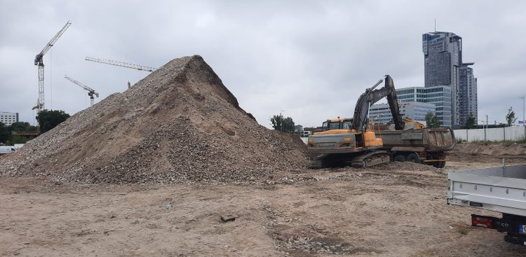 Plac budowy kompleksu Nova Marina w Gdyni. Na pierwszym planie, po lewej stronie, dominuje hałda piachu lub gruzu. Na prawo stoi żółta koparka gąsienicowa, która ładuje materiał na wywrotkę stojącą obok. Na ziemi widać ślady prac ziemnych i nieregularną, piaszczystą powierzchnię. W tle, po lewej stronie, widoczne są trzy żurawie budowlane, sugerujące dalsze prace konstrukcyjne w okolicy. Po prawej stronie, na horyzoncie, rysują się nowoczesne wieżowce, w tym jeden charakterystyczny, wysoki budynek o szklanej elewacji. Pomiędzy placem budowy a wieżowcami widać fragmenty innych budynków i miejskiej infrastruktury i zachmurzone