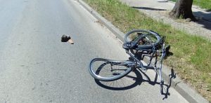 Zniszczony rower leży na drodze po zderzeniu z samochodem. Widać powyginane koła i ramę, na drodze leży jakiś element - prawdopodobnie urwana część któregoś z pojazdów lub but
