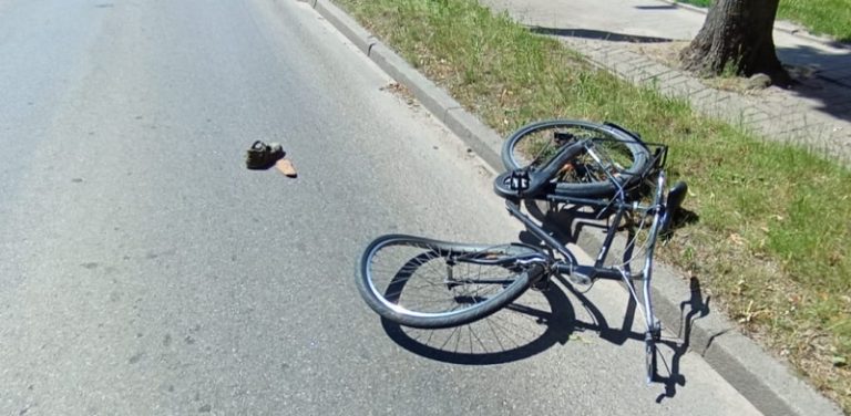 Zniszczony rower leży na drodze po zderzeniu z samochodem. Widać powyginane koła i ramę, na drodze leży jakiś element - prawdopodobnie urwana część któregoś z pojazdów lub but