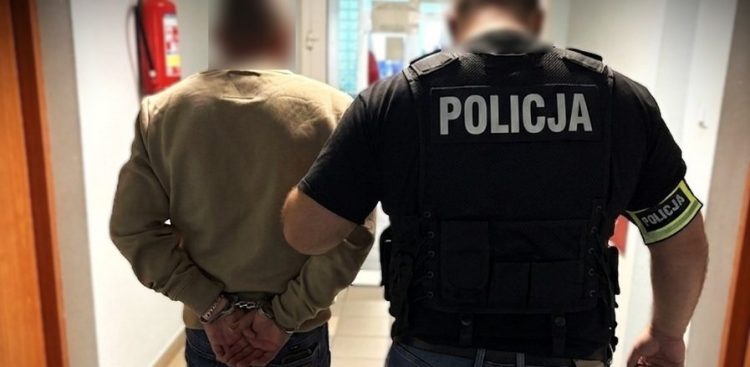 Po lewej zatrzymany z rękami skutymi kajdankami, po prawej prowadzący go rosły policjant. Postacie widziane od tyłu