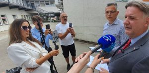 Konferencja posła PiS Michała Kowalskiego ws. rzekomych nieprawidłowości w Chojnickiej Spółdzielni Mieszkaniowej. Plenerowe spotkanie prasowe przed szarym, betonowym budynkiem. Po prawej stronie stoją dwaj mężczyźni – jeden w szarym garniturze i czerwonym krawacie przemawia, drugi w jasnej koszuli w paski i okularach słucha go, trzymając w rękach dokumenty. Przed nimi widoczne są dłonie dziennikarzy trzymające mikrofony z logo „Weekend FM” oraz „Radio Gdańsk”. Po lewej stronie stoją trzy osoby nagrywające wypowiedź telefonami komórkowymi, w tym kobieta w białej marynarce i ciemnych okularach przeciwsłonecznych korzystająca z gimbala.