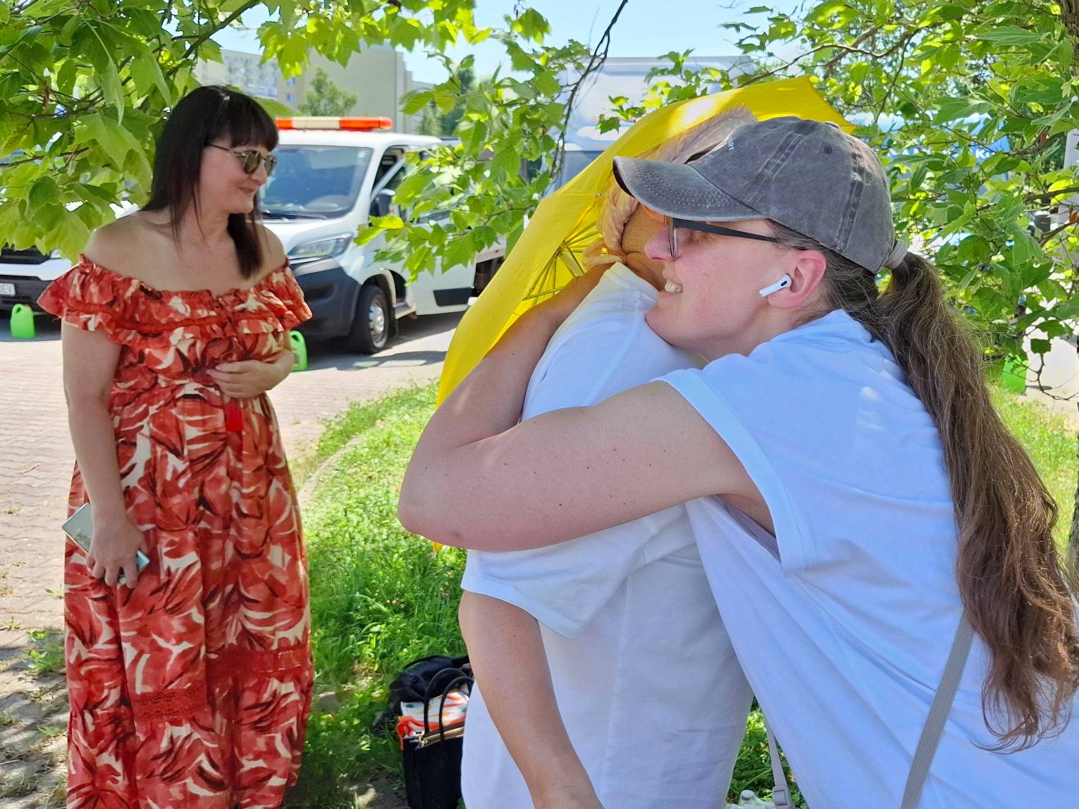 Pożegnanie wolontariuszy przed wyjazdem na Ukrainę konwoju pomocowego fundacji Zupa na Monciaku. Zdjęcie przedstawia scenę serdecznego pożegnania na świeżym powietrzu w słoneczny dzień. Na pierwszym planie po prawej stronie widoczne są dwie przytulające się osoby – kobieta w okularach, szarej czapce z daszkiem i białym T-shircie mocno obejmuje drugą osobę, nad którą widoczny jest fragment żółtego materiału. Po lewej stronie, w cieniu zielonych liści drzewa, stoi kobieta w długiej, czerwono-pomarańczowej wzorzystej sukience i okularach przeciwsłonecznych, która z uśmiechem przygląda się uściskowi. W tle zaparkowany jest biały samochód dostawczy z pomarańczowym światłem ostrzegawczym na dachu.
