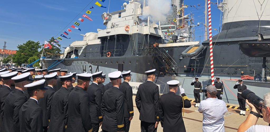 Uroczystość na nabrzeżu przy okręcie ORP „Błyskawica” (widoczny numer burtowy H34) w Gdyni. Po lewej stronie i na pierwszym planie stoi tyłem grupa oficerów Marynarki Wojennej w ciemnych mundurach i białych czapkach garnizonowych. W tle, nad pokładem okrętu, unosi się biały dym po wystrzale z salutu armatniego. Po prawej stronie widać salutujących żołnierzy oraz obserwujących wydarzenie cywilów, z których jeden fotografuje uroczystość nadania promocji oficerskich