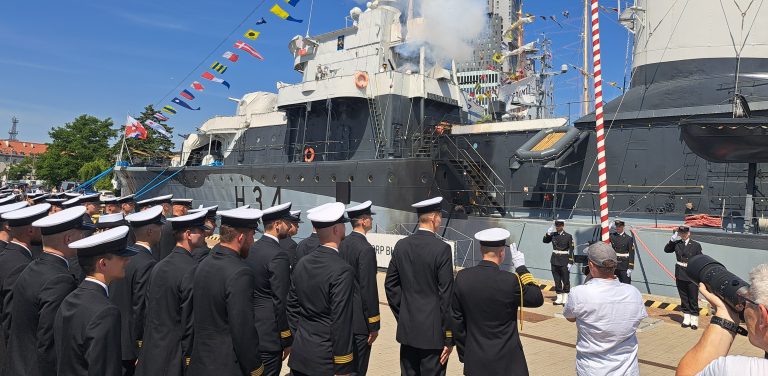 Uroczystość na nabrzeżu przy okręcie ORP „Błyskawica” (widoczny numer burtowy H34) w Gdyni. Po lewej stronie i na pierwszym planie stoi tyłem grupa oficerów Marynarki Wojennej w ciemnych mundurach i białych czapkach garnizonowych. W tle, nad pokładem okrętu, unosi się biały dym po wystrzale z salutu armatniego. Po prawej stronie widać salutujących żołnierzy oraz obserwujących wydarzenie cywilów, z których jeden fotografuje uroczystość nadania promocji oficerskich