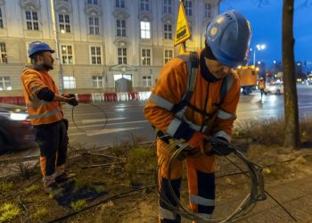 Dwaj robotnicy w pomarańczowych strojach odblaskowych i niebieskich kaskach pracują o zmierzchu na pasie zieleni przy ulicy 3 Maja w Gdańsku. Mężczyzna na pierwszym planie zwija grubą stalową linę, a drugi robotnik w tle trzyma przewód. W tle widoczna jest jezdnia z jadącymi samochodami, klasyczny gmach budynku oraz żółty znak ostrzegawczy z tramwajem. Zdjęcie ilustruje utrudnienia dla pasażerów z powodu remontu Węzła Hucisko - zmiany rozkłady jazdy tramwajów i autobusów w Gdańsku od soboty, 27 czerwca