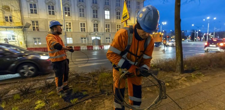 Dwaj robotnicy w pomarańczowych strojach odblaskowych i niebieskich kaskach pracują o zmierzchu na pasie zieleni przy ulicy 3 Maja w Gdańsku. Mężczyzna na pierwszym planie zwija grubą stalową linę, a drugi robotnik w tle trzyma przewód. W tle widoczna jest jezdnia z jadącymi samochodami, klasyczny gmach budynku oraz żółty znak ostrzegawczy z tramwajem. Zdjęcie ilustruje utrudnienia dla pasażerów z powodu remontu Węzła Hucisko - zmiany rozkłady jazdy tramwajów i autobusów w Gdańsku od soboty, 27 czerwca