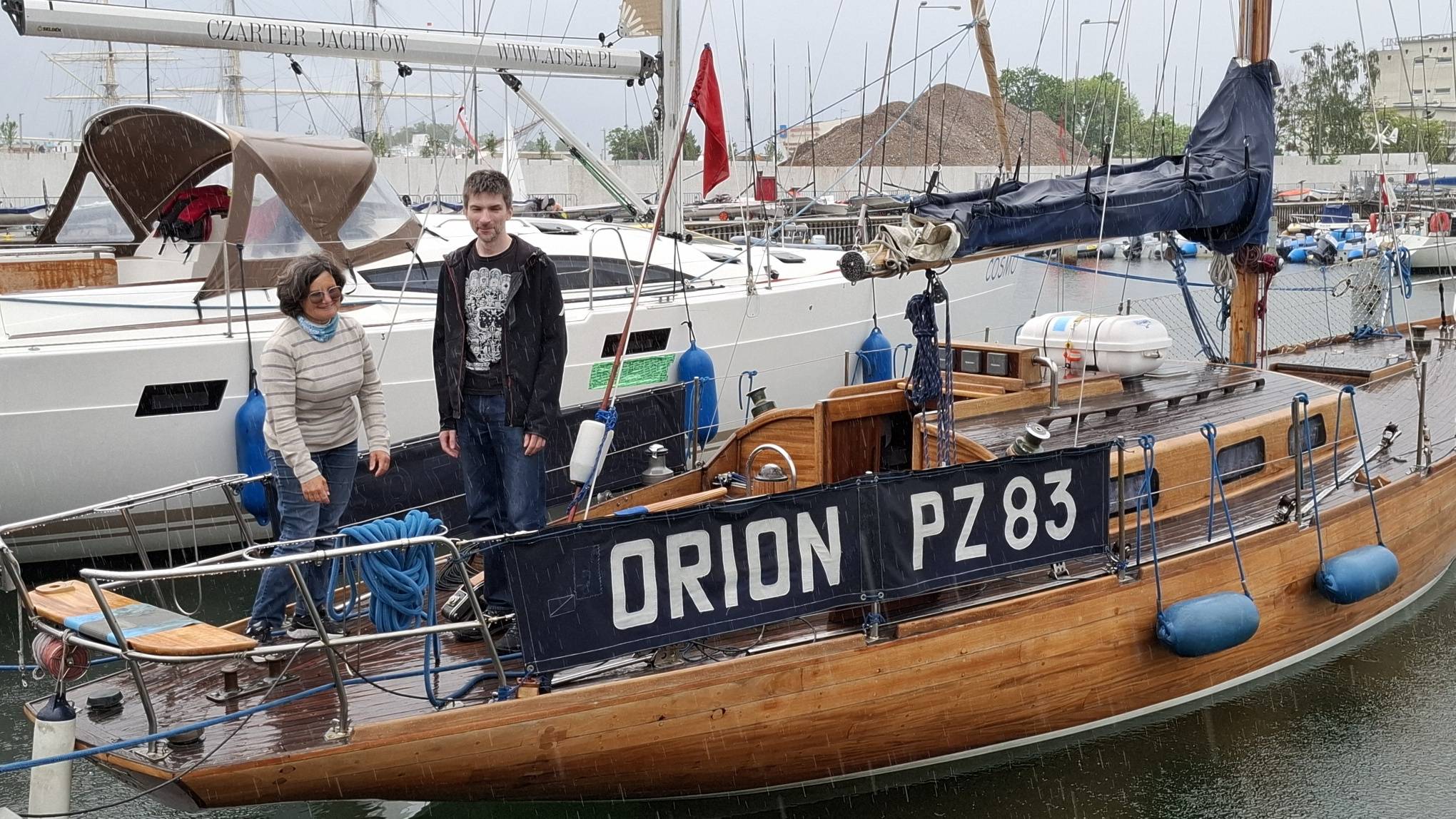 Drewniany jacht s/y Orion (PZ 83) stojący w marinie z dwoma osobami stojącymi na pokładzie. W tle widać inne jachty