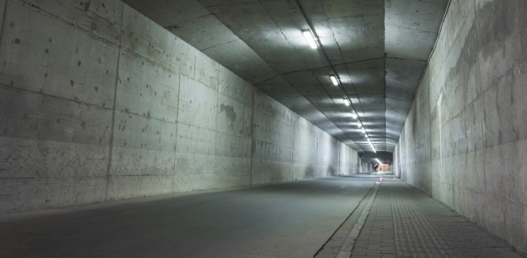 Długi, betonowy tunel (schron) z rzędem jasnych świateł na suficie, widoczny od strony wejścia. Droga jest pusta, a po prawej stronie znajduje się chodnik
