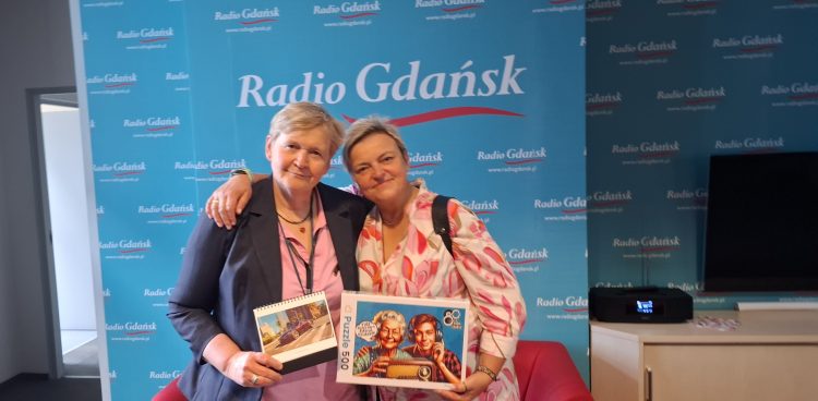 Dwie uśmiechnięte kobiety w średnim wieku stoją obok siebie, obejmując się ramionami, przed ścianą z logo „Radio Gdańsk”. Kobieta po lewej to redaktor Anna Rębas - ma krótkie, jasne włosy, ubrana jest w marynarkę w ciemnym kolorze, jasną koszulę i naszyjnik. W lewej ręce trzyma kalendarz z obrazkiem. Kobieta po prawej to dr Dagmara Wiewiórkowska-Garczewska - również ma krótkie jasne włosy, ubrana jest w kolorową bluzkę z wzorami. W prawej ręce trzyma pudełko z puzzlami, na którym widnieje miniatura muralu na 80-lecie radia Gdańsk (postaci starszej kobiety i chłopaka, być może wnuka, którzy słuchają radia). Obie kobiety wydają się być w dobrym nastroju. Tło stanowi niebieska ściana z powtarzającym się logo „Radio Gdańsk” w białym kolorze