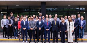 Grupa około 30 osób stojących przed nowoczesnym budynkiem z dużymi szklanymi oknami i ciemną fasadą. Większość osób jest ubrana w stroje biznesowe lub smart casual. W centralnej części zdjęcia znajduje się rząd mężczyzn w garniturach. Wokół nich stoją inni uczestnicy, w tym kilka kobiet. Z tyłu widać również młodsze osoby. Wszyscy patrzą w stronę obiektywu, a na ich twarzach widać uśmiechy lub neutralne wyrazy twarzy. To zdjęcie pamiątkowe przedstawicieli pięciu gdańskich uczelni, które podpisały wspólne stanowisko popierające ustawę o związku metropolitalnym w województwie pomorskim