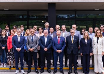 Grupa około 30 osób stojących przed nowoczesnym budynkiem z dużymi szklanymi oknami i ciemną fasadą. Większość osób jest ubrana w stroje biznesowe lub smart casual. W centralnej części zdjęcia znajduje się rząd mężczyzn w garniturach. Wokół nich stoją inni uczestnicy, w tym kilka kobiet. Z tyłu widać również młodsze osoby. Wszyscy patrzą w stronę obiektywu, a na ich twarzach widać uśmiechy lub neutralne wyrazy twarzy. To zdjęcie pamiątkowe przedstawicieli pięciu gdańskich uczelni, które podpisały wspólne stanowisko popierające ustawę o związku metropolitalnym w województwie pomorskim