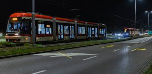 Trzy czerwono-białe tramwaje jadące nocą. Pierwszy tramwaj, z jasno oświetlonymi wnętrzami, jest widoczny z boku. W tle po prawej stronie widać kolejne dwa zbliżające się. Ulica jest oświetlona latarniami, a na asfalcie widoczne są żółte strzałki kierunkowe