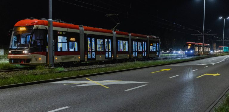 Trzy czerwono-białe tramwaje jadące nocą. Pierwszy tramwaj, z jasno oświetlonymi wnętrzami, jest widoczny z boku. W tle po prawej stronie widać kolejne dwa zbliżające się. Ulica jest oświetlona latarniami, a na asfalcie widoczne są żółte strzałki kierunkowe