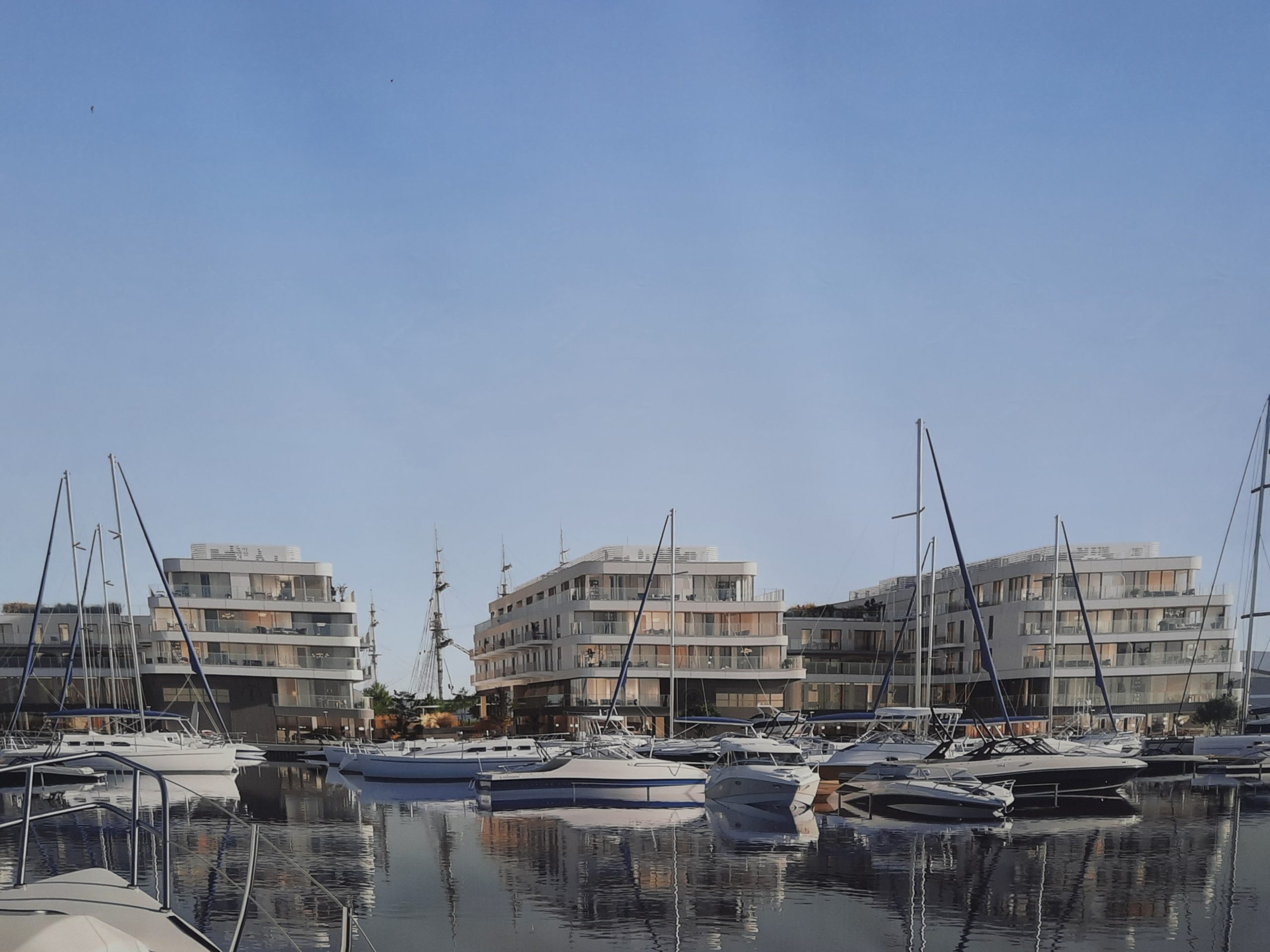 Wizualizacja kompleksu Nova Marina w Gdyni przedstawia marinę z nowoczesnymi budynkami mieszkalnymi. Na pierwszym planie znajduje się spokojna woda zatoki, w której odbijają się jachty i otoczenie, tworząc symetryczny obraz. Liczne białe jachty i motorówki o różnych rozmiarach, które są zacumowane przy marinie