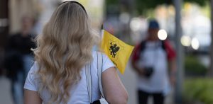 Kobieta o długich, falowanych blond włosach, widziana od tyłu, idzie miejską ulicą i trzyma w dłoni małą żółtą flagę z czarnym gryfem, który jest symbolem Kaszub. Tło z innymi przechodniami jest rozmyte