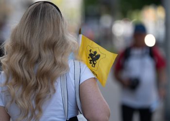 Kobieta o długich, falowanych blond włosach, widziana od tyłu, idzie miejską ulicą i trzyma w dłoni małą żółtą flagę z czarnym gryfem, który jest symbolem Kaszub. Tło z innymi przechodniami jest rozmyte