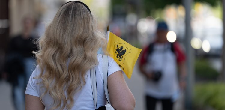 Kobieta o długich, falowanych blond włosach, widziana od tyłu, idzie miejską ulicą i trzyma w dłoni małą żółtą flagę z czarnym gryfem, który jest symbolem Kaszub. Tło z innymi przechodniami jest rozmyte
