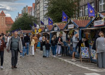 Szeroka brukowana ulica w Gdańsku podczas Jarmarku św. Dominika. Po prawej stronie ciągnie się rząd drewnianych domków handlowych ozdobionych flagami z kolorowym kogutem – symbolem jarmarku. Na jednym z szyldów widnieje napis „Tradycyjne wyroby z Podlasia”. Ulicą spacerują ludzie, w tym para na pierwszym planie. W tle wznosi się zabytkowy, neogotycki budynek Hali Targowej z czerwonej cegły