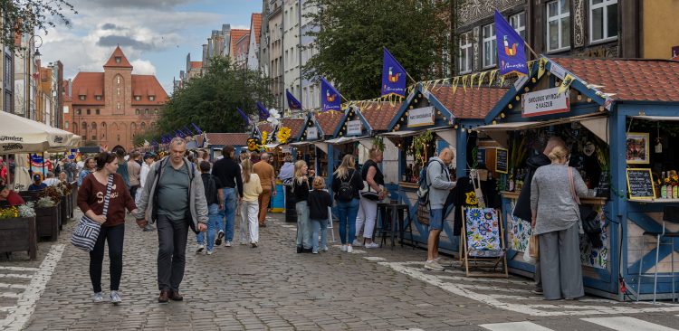 Szeroka brukowana ulica w Gdańsku podczas Jarmarku św. Dominika. Po prawej stronie ciągnie się rząd drewnianych domków handlowych ozdobionych flagami z kolorowym kogutem – symbolem jarmarku. Na jednym z szyldów widnieje napis „Tradycyjne wyroby z Podlasia”. Ulicą spacerują ludzie, w tym para na pierwszym planie. W tle wznosi się zabytkowy, neogotycki budynek Hali Targowej z czerwonej cegły