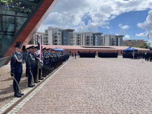 Po lewej stronie kadru stoi kompania honorowa policji w galowych mundurach z białymi sznurami naramiennymi, trzymająca sztandar oraz broń reprezentacyjną. Ponad nimi, po lewej stronie, wznosi się fragment charakterystycznej, pochylonej, czerwonawej elewacji Muzeum II Wojny Światowej. Na rozległym, brukowanym placu w tle stoją dwa duże, uformowane w rzędach pododdziały policji w ciemnych mundurach. W oddali, za niskim murkiem, widoczne są nowoczesne, jasne bloki mieszkalne pod częściowo zachmurzonym niebem. Pomorscy policjanci świętowali 107. rocznicę powstania policji państwowej