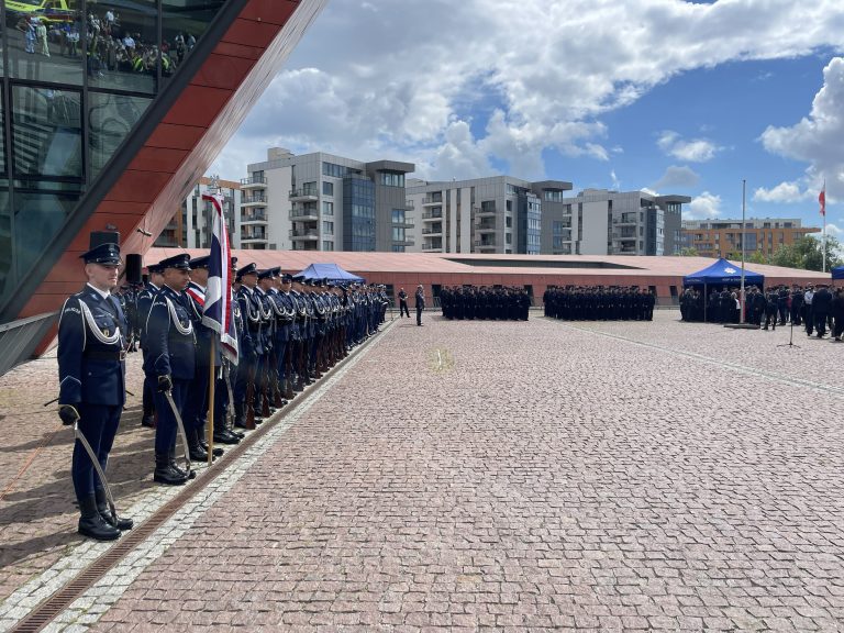 Po lewej stronie kadru stoi kompania honorowa policji w galowych mundurach z białymi sznurami naramiennymi, trzymająca sztandar oraz broń reprezentacyjną. Ponad nimi, po lewej stronie, wznosi się fragment charakterystycznej, pochylonej, czerwonawej elewacji Muzeum II Wojny Światowej. Na rozległym, brukowanym placu w tle stoją dwa duże, uformowane w rzędach pododdziały policji w ciemnych mundurach. W oddali, za niskim murkiem, widoczne są nowoczesne, jasne bloki mieszkalne pod częściowo zachmurzonym niebem. Pomorscy policjanci świętowali 107. rocznicę powstania policji państwowej