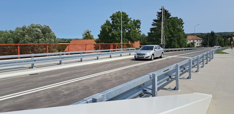 Srebrny samochód osobowy jadący po nowym moście drogowym w Nowakowie. Jezdnię z podwójną linią ciągłą oddzielają od boków metalowe bariery ochronne, a po lewej stronie widoczna jest pomarańczowo-czerwona balustrada zabezpieczająca chodnik. W tle znajdują się zielone drzewa, fragmenty zabudowań oraz bezchmurne, jasnoniebieskie niebo