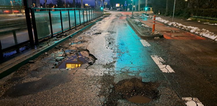 Głębokie wyrwy i dziury z kałużami wody na mocno zniszczonej jezdni ulicy Wita Stwosza w Gdańsku, tuż obok przystanku tramwajowego po zmroku. Ul. Wita Stwosza w Gdańsku-Oliwie doczeka się odnowy nawierzchni