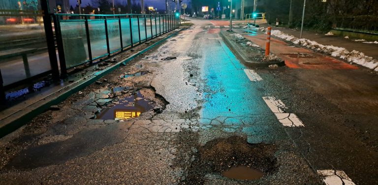 Głębokie wyrwy i dziury z kałużami wody na mocno zniszczonej jezdni ulicy Wita Stwosza w Gdańsku, tuż obok przystanku tramwajowego po zmroku. Ul. Wita Stwosza w Gdańsku-Oliwie doczeka się odnowy nawierzchni