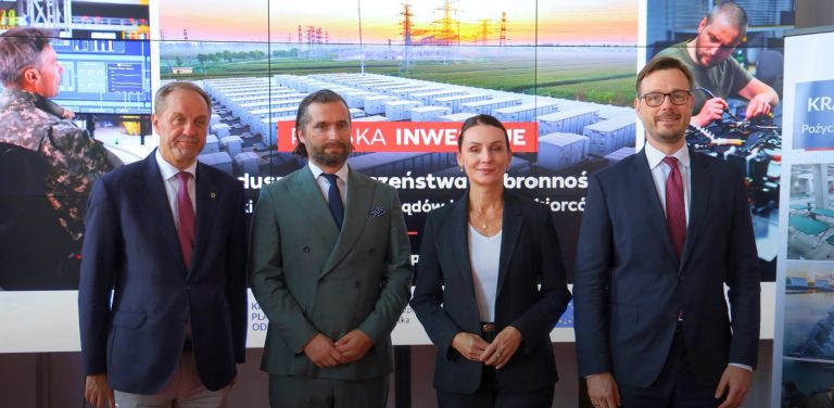 Cztery osoby w eleganckich strojach biznesowych stoją obok siebie w rzędzie na tle dużego ekranu multimedialnego. Od lewej: mężczyzna w granatowej marynarce i różowym krawacie, brodaty mężczyzna w zielonym garniturze dwurzędowym, kobieta w ciemnej marynarce i białym topie oraz mężczyzna w okularach, ciemnym garniturze i bordowym krawacie. Na ekranie w tle widoczny jest czerwony baner z napisem „POLSKA INWESTUJE” oraz grafika przedstawiająca instalację przemysłową z rzędami kontenerów i słupami energetycznymi