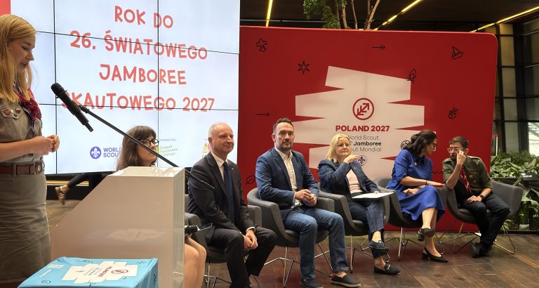 Konferencja prasowa na tle ekranu z napisem „ROK DO 26. ŚWIATOWEGO JAMBOREE SKAUTOWEGO 2027”. Po lewej stronie harcerka przemawia przy mikrofonie, a po prawej siedzą uczestnicy panelu – przedstawiciele władz, Kancelarii Prezydenta RP i organizatorów na tle czerwonej ścianki z logo „Poland 2027”
