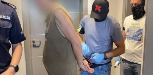 Policjanci w rękawiczkach ochronnych i mężczyzna w kajdankach z zamazaną twarzą, ubrany w bezrękawnik. Po lewej stronie stoi umundurowany policjant, a po prawej drugi cywilny funkcjonariusz z zasłoniętą twarzą. Zatrzymany zaatakował w Rumi nożem innego mężczyznę na tle narodowościowym