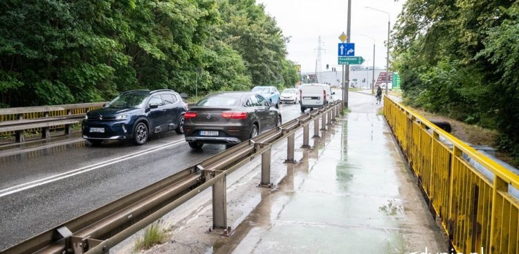Mokra jezdnia na wiadukcie przy ul. płk. Dąbka w Gdyni, po której jadą samochody osobowe. Po prawej stronie, za metalową barierą energochłonną, przebiega chodnik zabezpieczony żółtą balustradą. W tle widoczne są drzewa oraz niebieski znak drogowy wskazujący kierunki na Kosakowo i Rewę